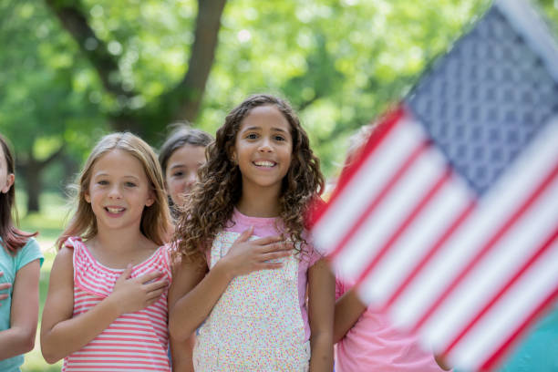 Diverse group of elementary age school girls are standing outdoors with hands over hearts. Children are smiling and reciting the Pledge of Allegience to the American flag.